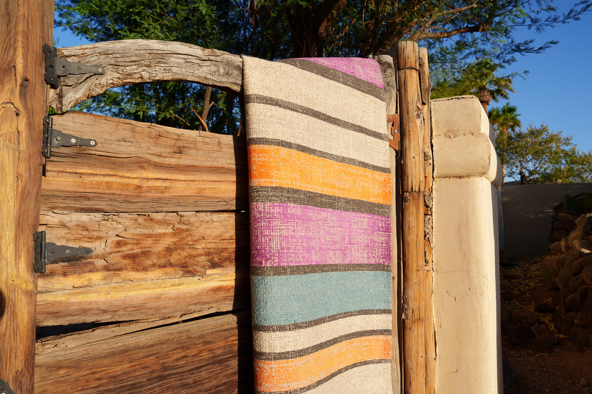 A multicolor striped Mother Ruggers rug draped over a wooden gate in warm sunlight.